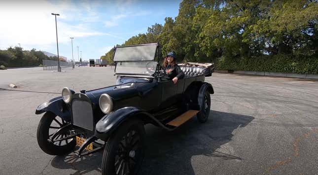 A curly haired woman in a bucket hat sits smugly in a black 1915 Dodge Bros Touring Car in a parking lot.
