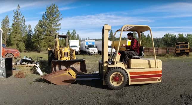 A man in flannel pilots a striped forklift in front of a bunch of old machinery