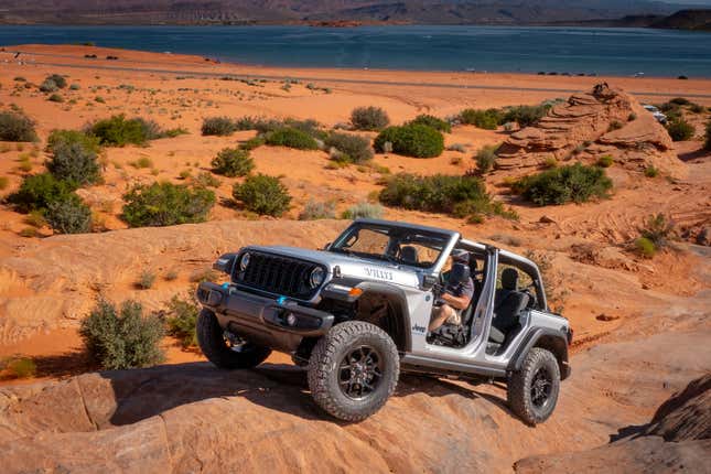 This photo shows a Jeep Wrangler hybrid with no roof or doors climbing an incline overlooking a lake.